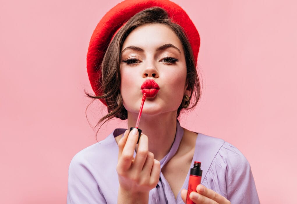 Portrait of young girl in red beret painting her lips with bright lipstick on pink background
