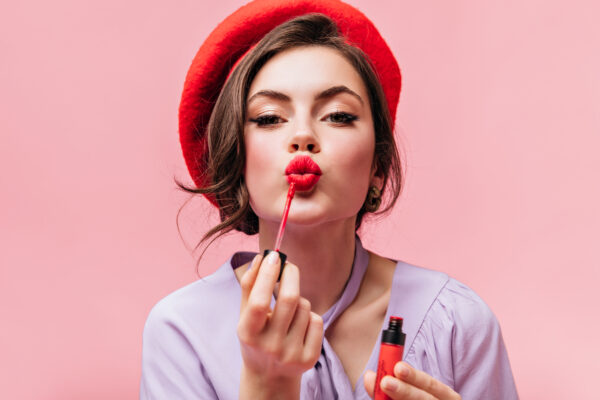 Portrait of young girl in red beret painting her lips with bright lipstick on pink background